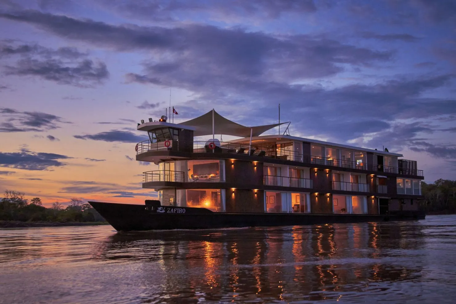 Crucero de lujo Zafiro en el río Amazonas al ocaso, con las cabinas iluminadas reflejadas en el agua